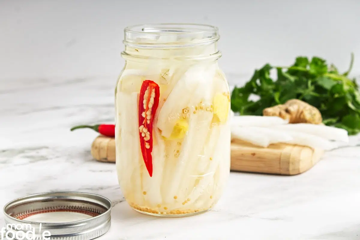 Side of of a jar with white radishes and a piece of red pepper.