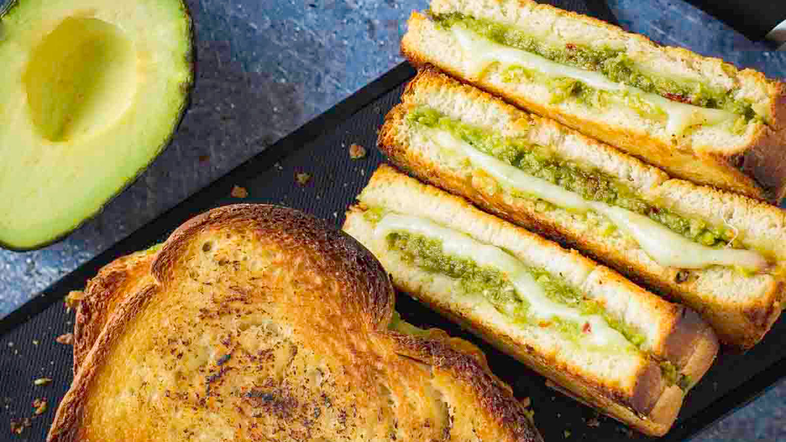 Grilled cheese with avocado cut in half standing on a black board.