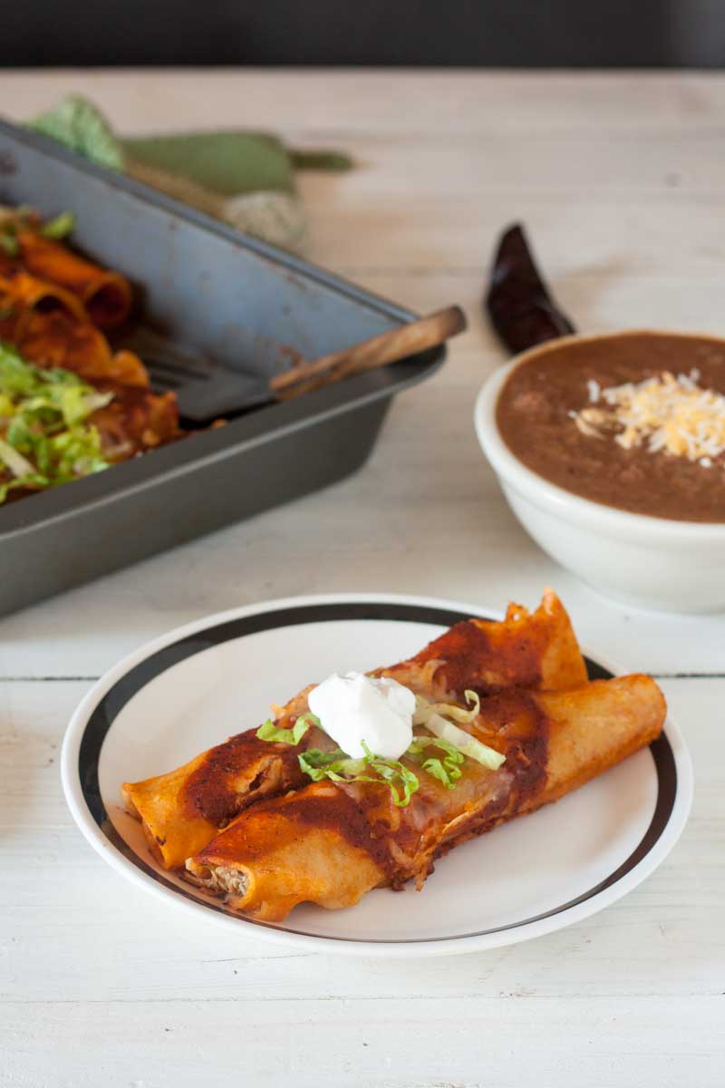 2 chicken enchiladas on white plate garnished with sour cream with refried beans