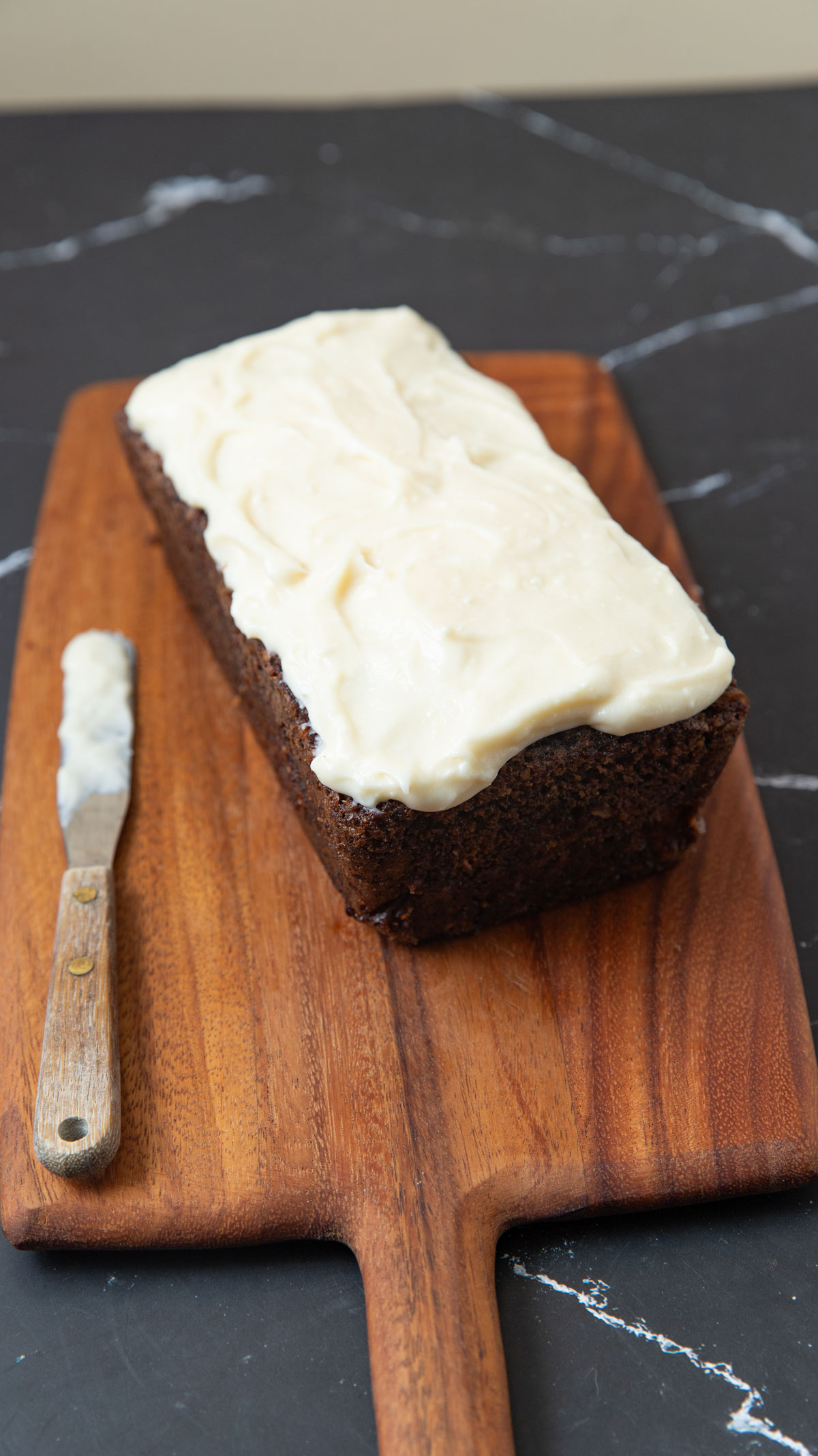 Whole carrot cake loaf on a wooden board with frosting knife on the board, too.