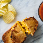 Grilled cheese sandwiches on a white board next to tomato soup and chips.