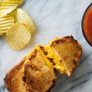 Grilled cheese sandwiches on a white board next to tomato soup and chips.
