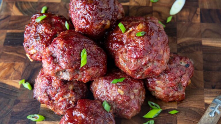 Smoked meatballs with BBQ sauce piled on a wooden cutting board and garnished with chopped greeen onion.