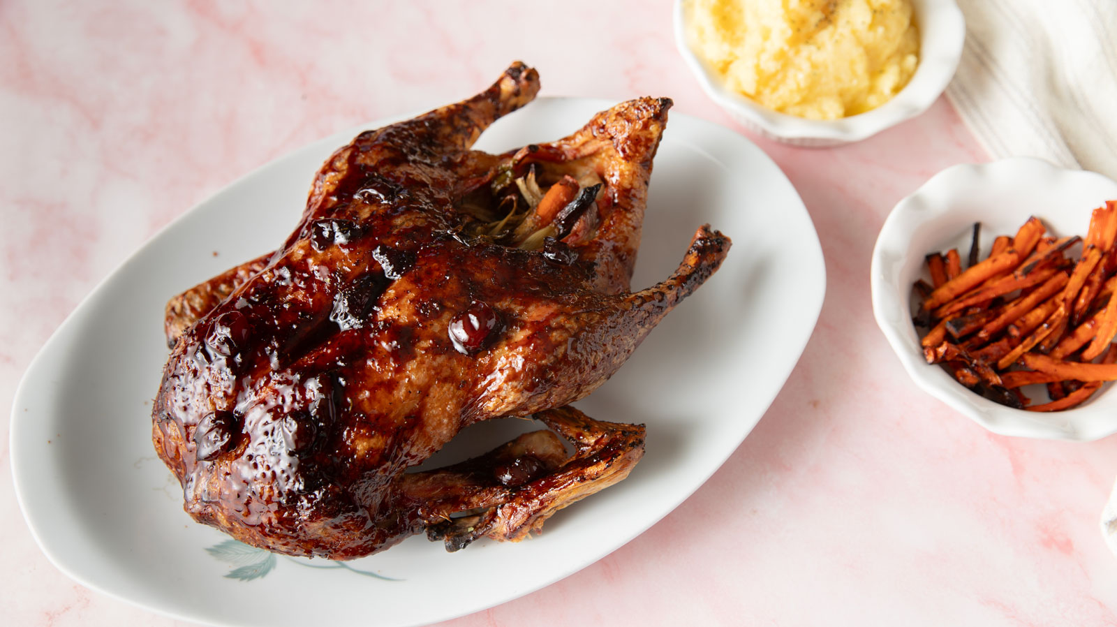 Glazed duck with visible cherries on a platter near bowls of carrots and mashed potatoes.