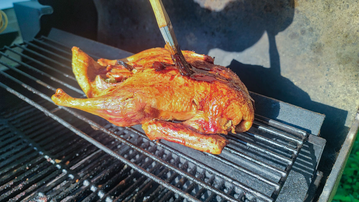 Whole duck on the grill while a hand brushes it with cherry glaze.