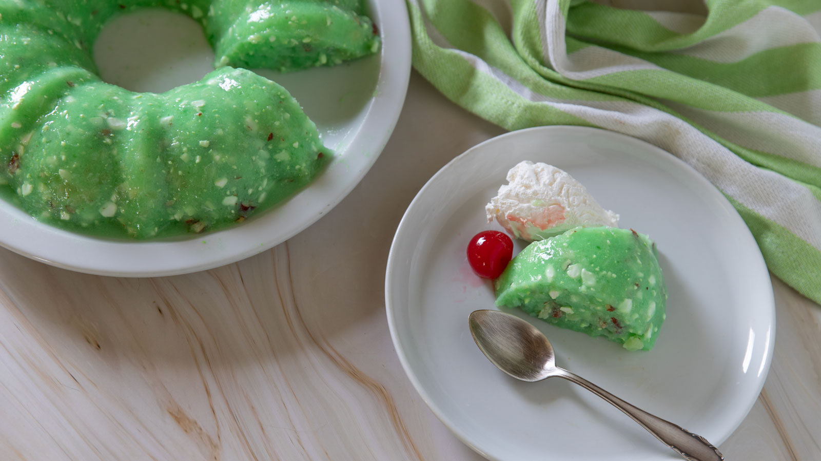 Serving of green jello salad on a white plate with the jello mold in the background.